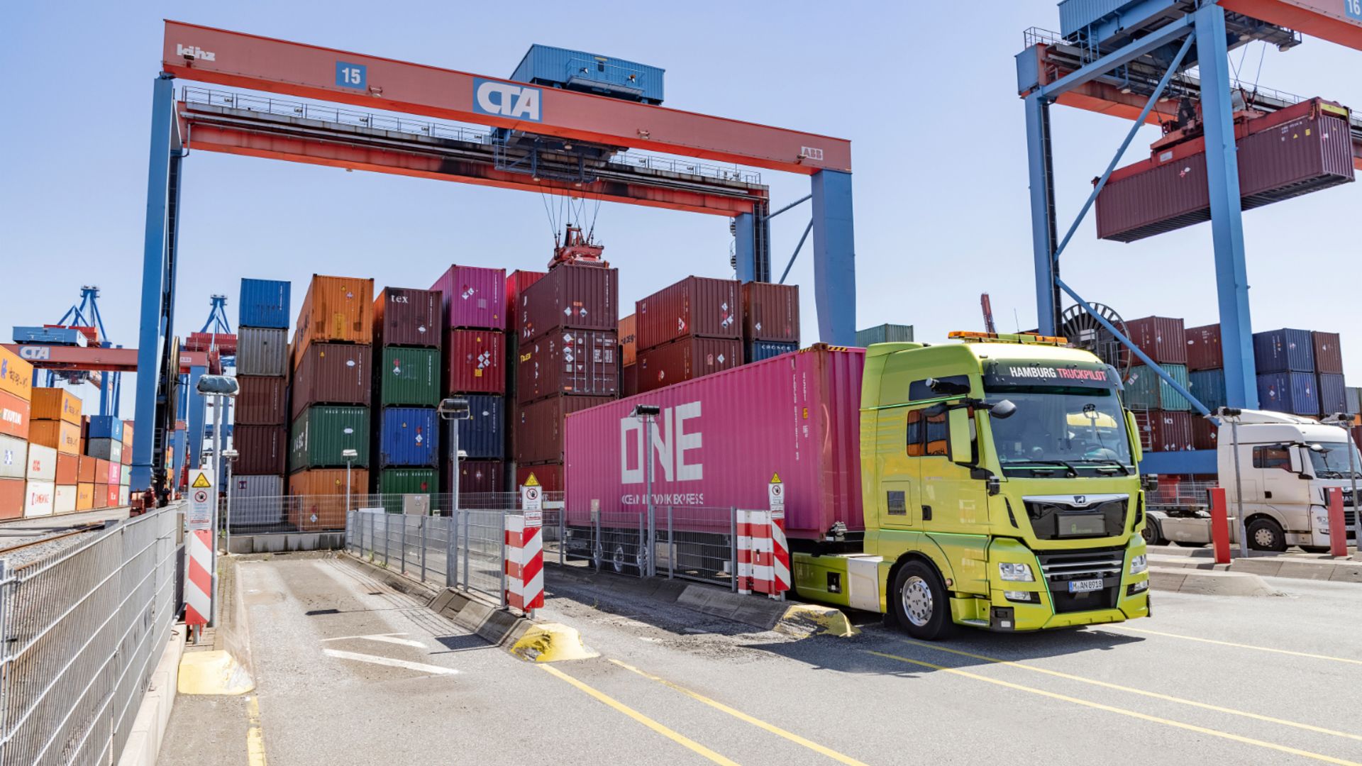 Yellow truck transports a pink container at the port, surrounded by stacked containers and cranes.