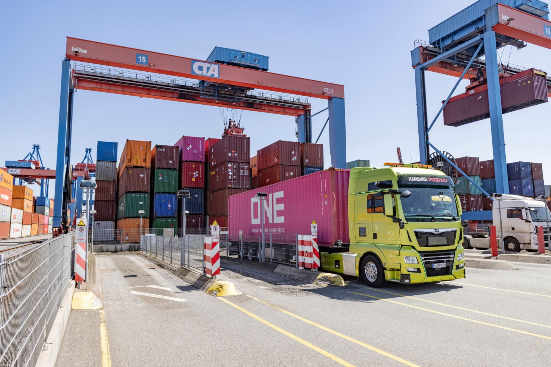 Yellow truck transports a pink container at the port, surrounded by stacked containers and cranes.
